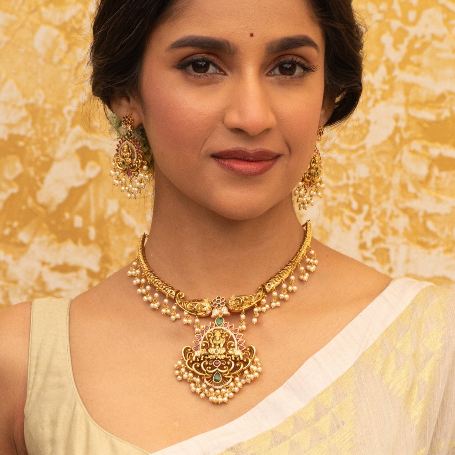 An image of a temple necklace set, featuring goddess Laxmi idols, adorned with white beads and red - green gemstones.